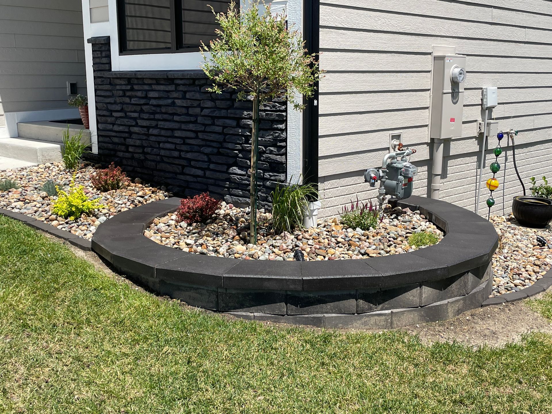 Curved stone flower bed with mulch, small tree, and gray house siding in front of a yard