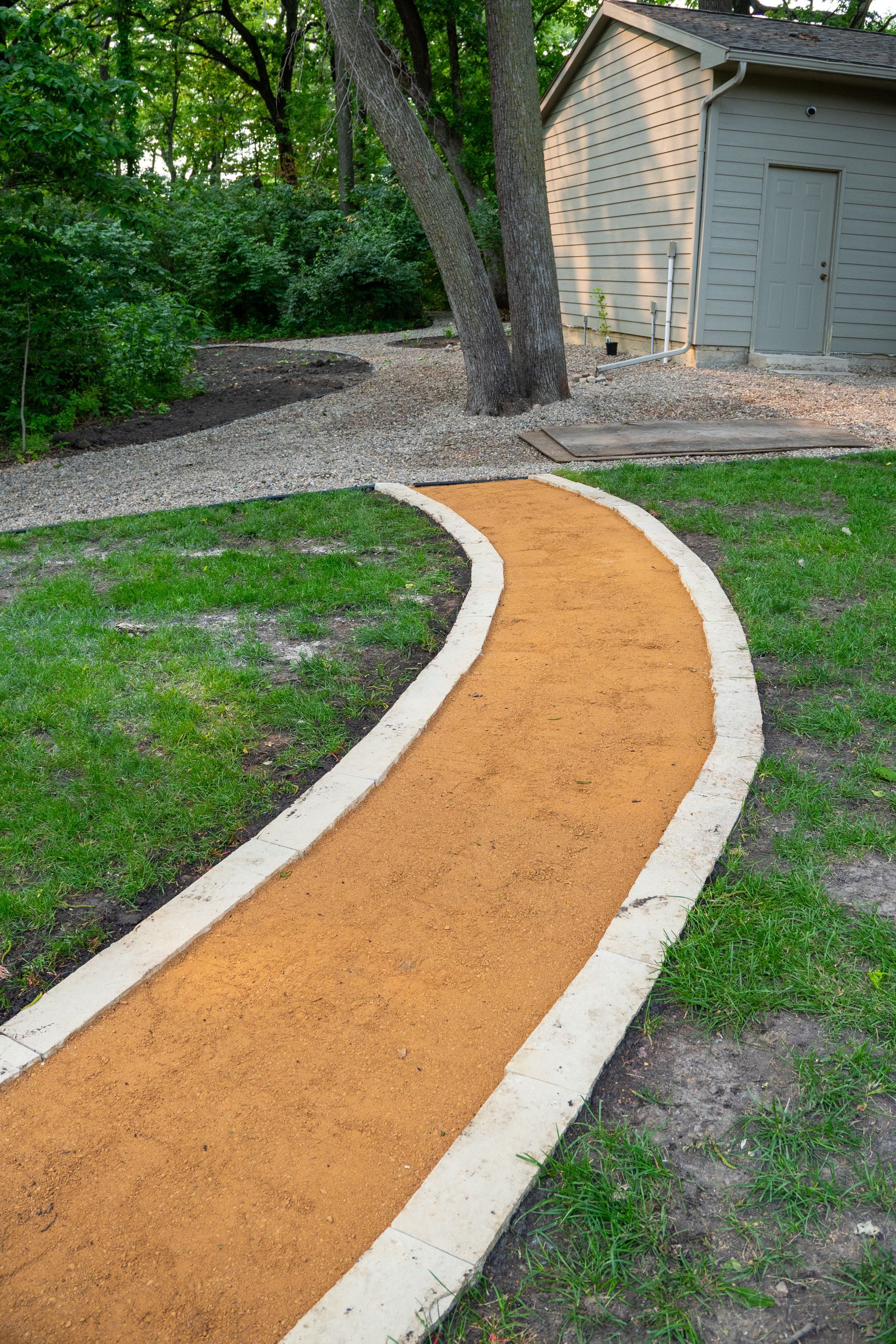 Curved orange walkway with white borders through a grassy yard beside trees and a shed
