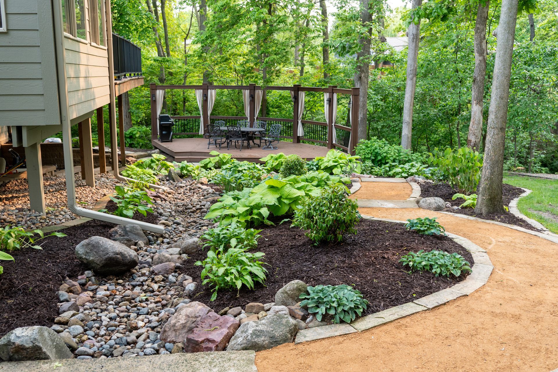 Landscaped garden with mulched beds, shrubs, stone edging, and a wooden deck in a wooded backyard