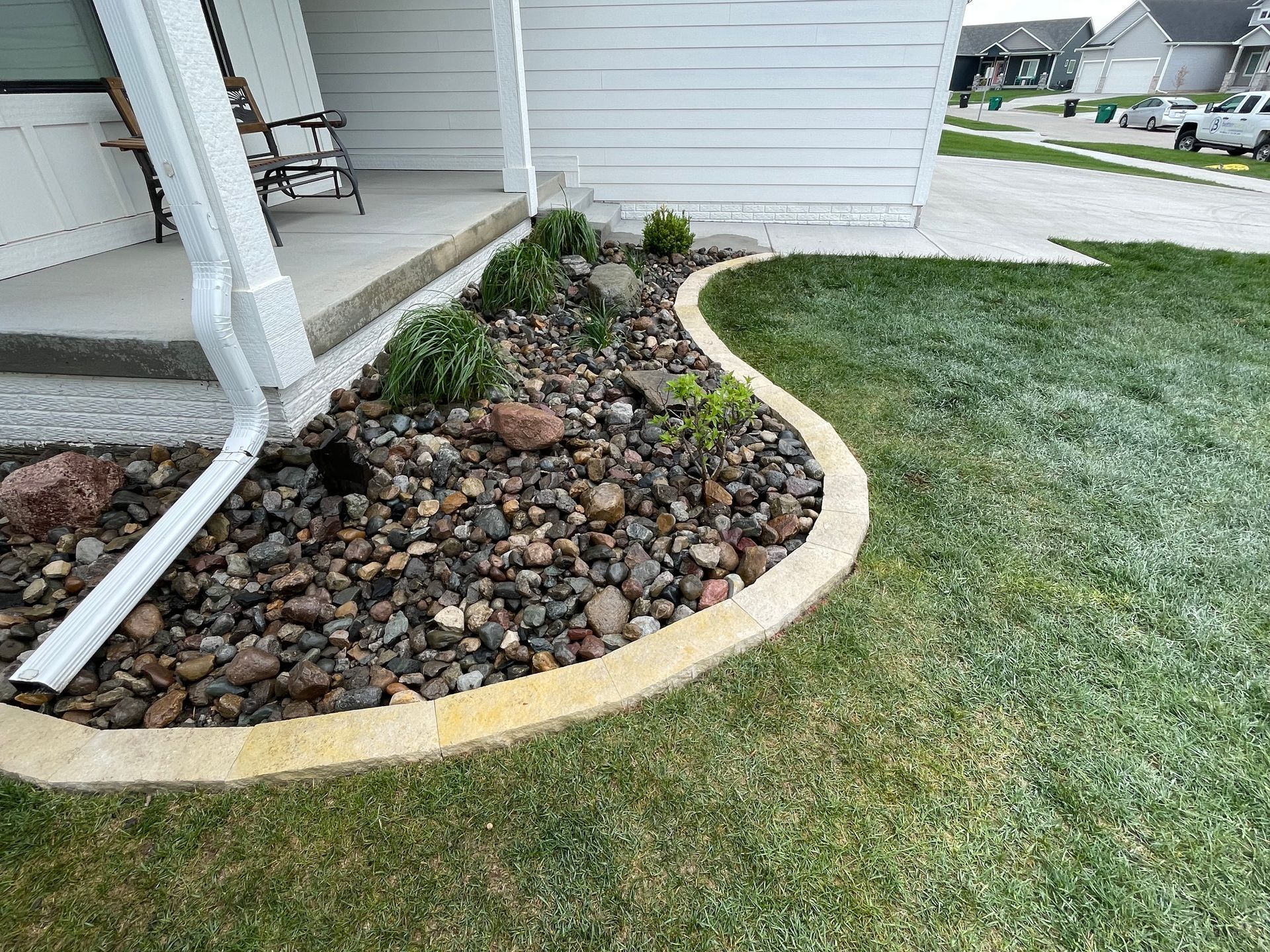 Curved rock garden bed beside a white house porch, bordered by grass and mulch with small shrubs.