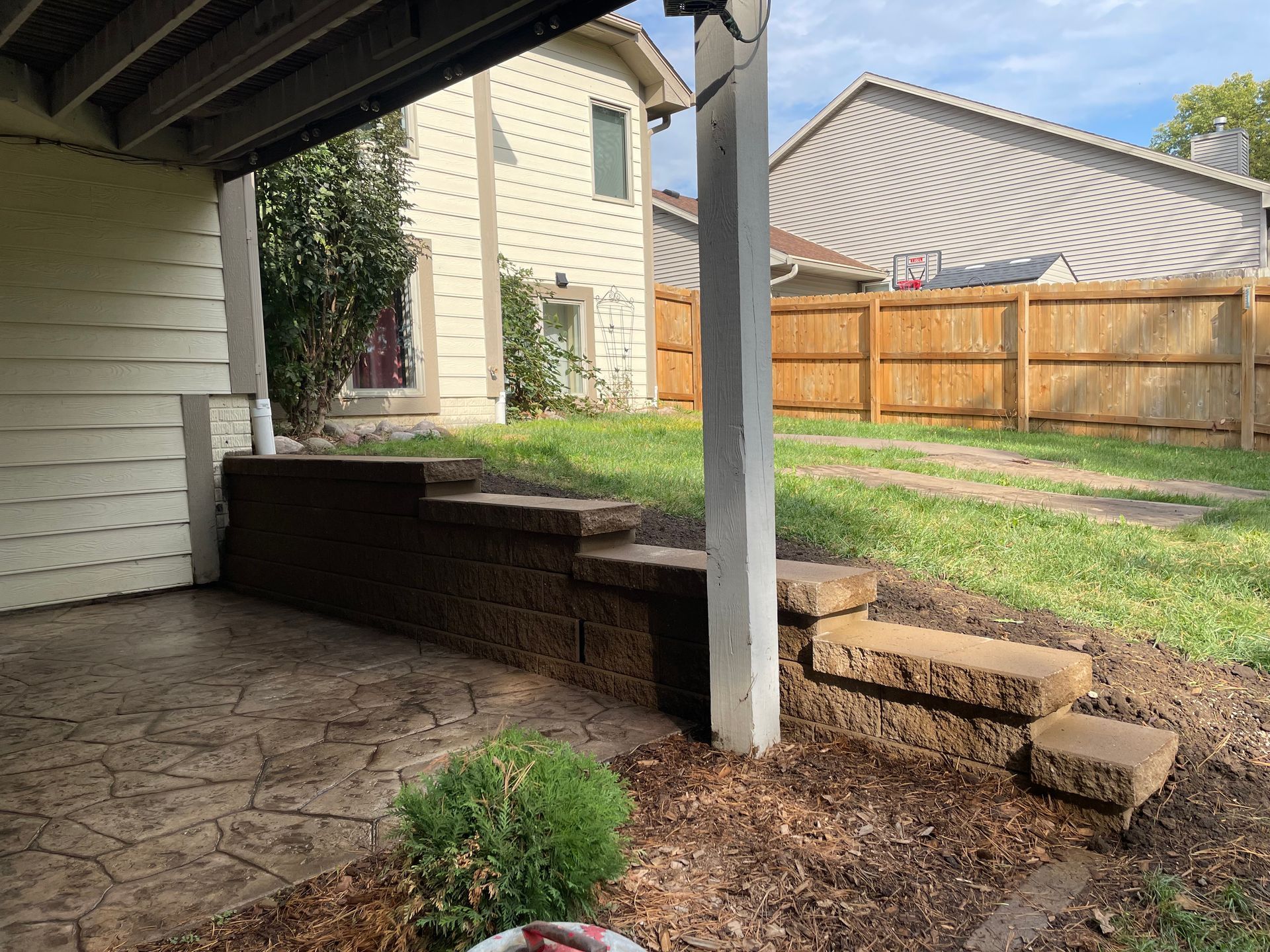 Covered backyard patio with stone floor, retaining wall, steps, white post, and fenced lawn under a blue sky