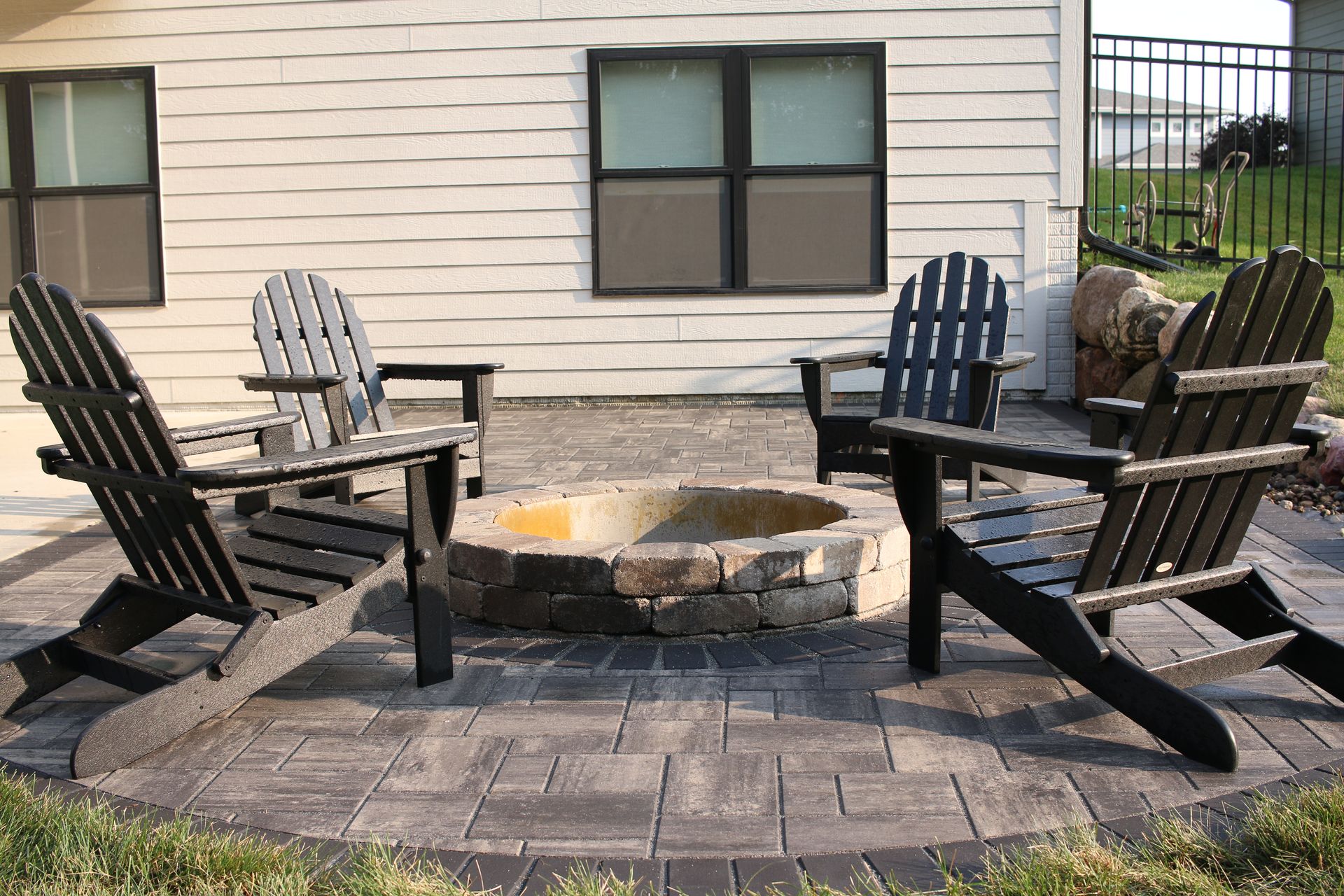 Four Adirondack chairs around a stone fire pit on a patio beside a house