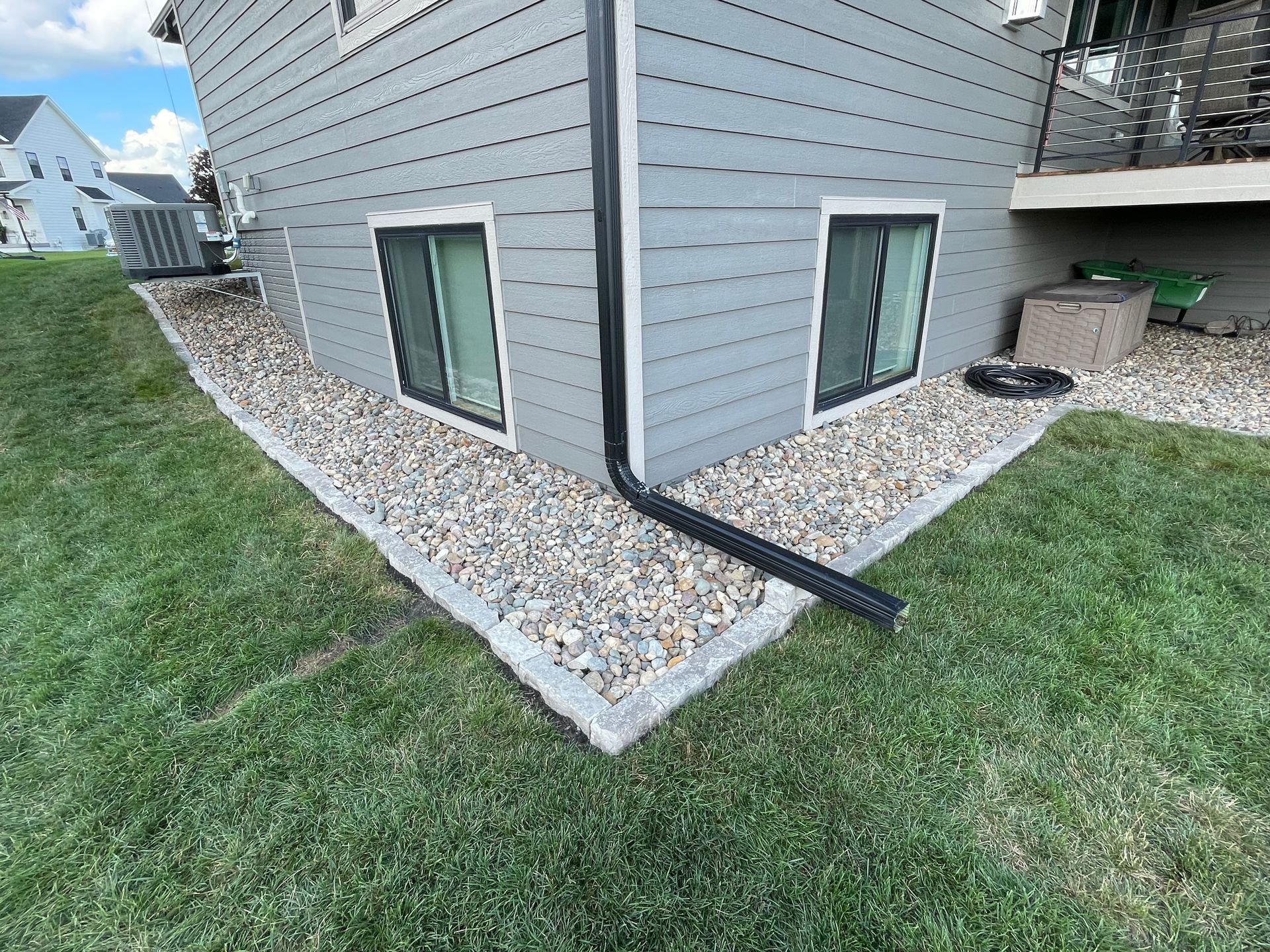 Corner of a gray house with basement windows, white gravel border, and a black downspout over green grass