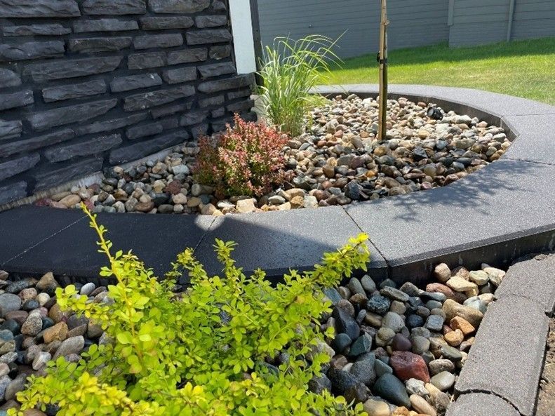 A modern landscaping with stone edging, decorative rocks, and small shrubs near a house wall Curved stone-edged garden bed with small rocks, young plants, and a dark retaining wall beside a driveway