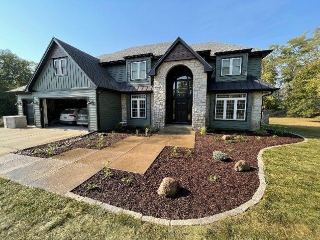 A front yard of a modern two-story house with fresh landscaping, mulch beds, and stone edging Large dark gray house with arched entry, garage, and landscaped front yard
