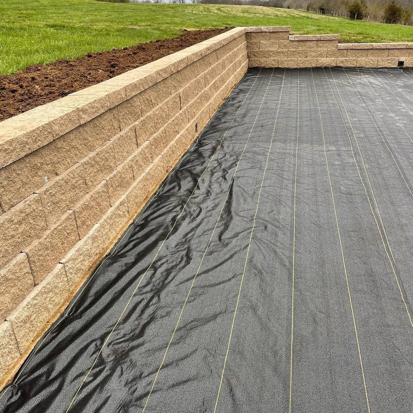 Tan retaining wall bordering a black landscape fabric-covered slope beside a grassy lawn