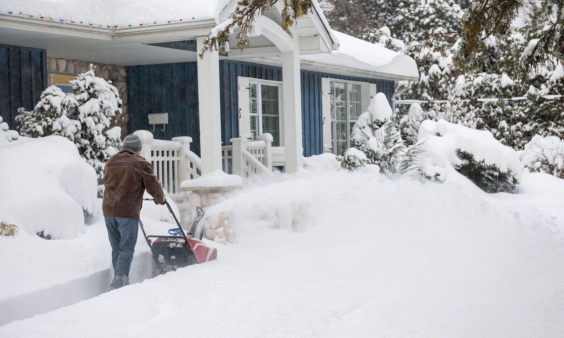 Person shoveling deep snow outside a house with snow-covered trees and yard