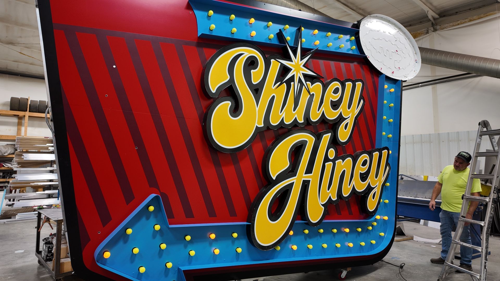 Colorful “Shine Hiney” sign with red stripes and blue lights, in a workshop beside two workers