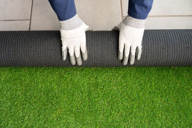 Hands wearing white work gloves unroll a length of green artificial turf on a light-colored tiled floor.