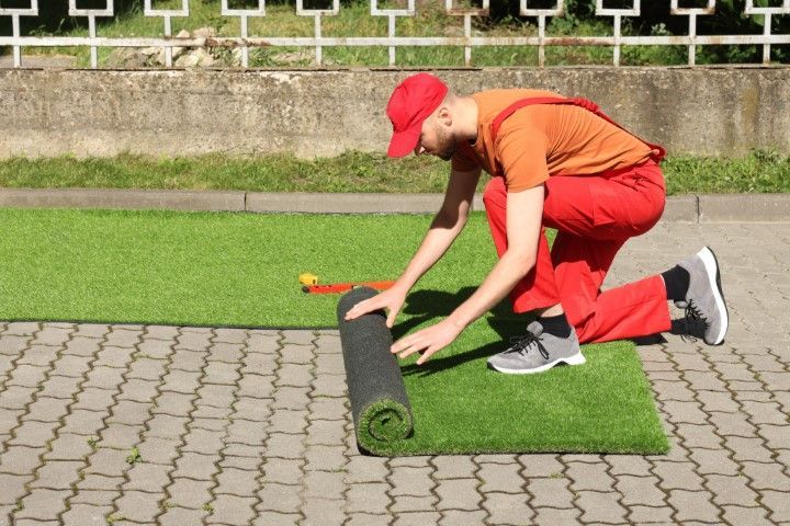 A person in red work clothes kneels on a paved surface, unrolling a strip of artificial grass.