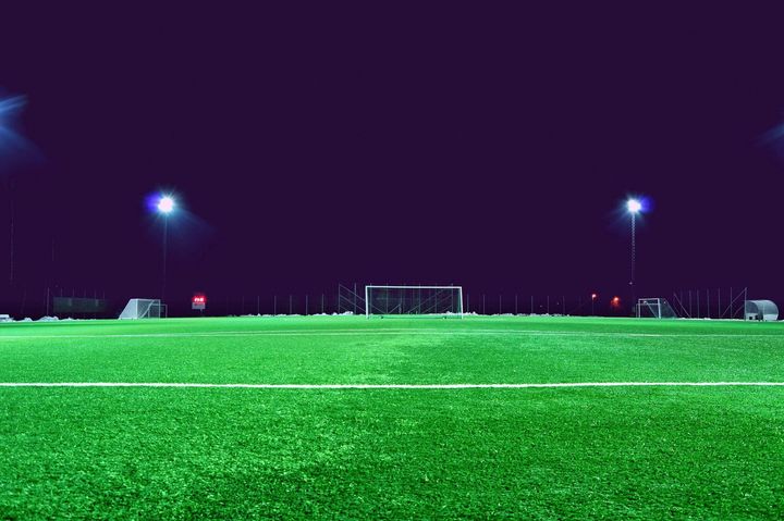 A soccer field at night, illuminated by bright floodlights, with a goalpost in the distance.