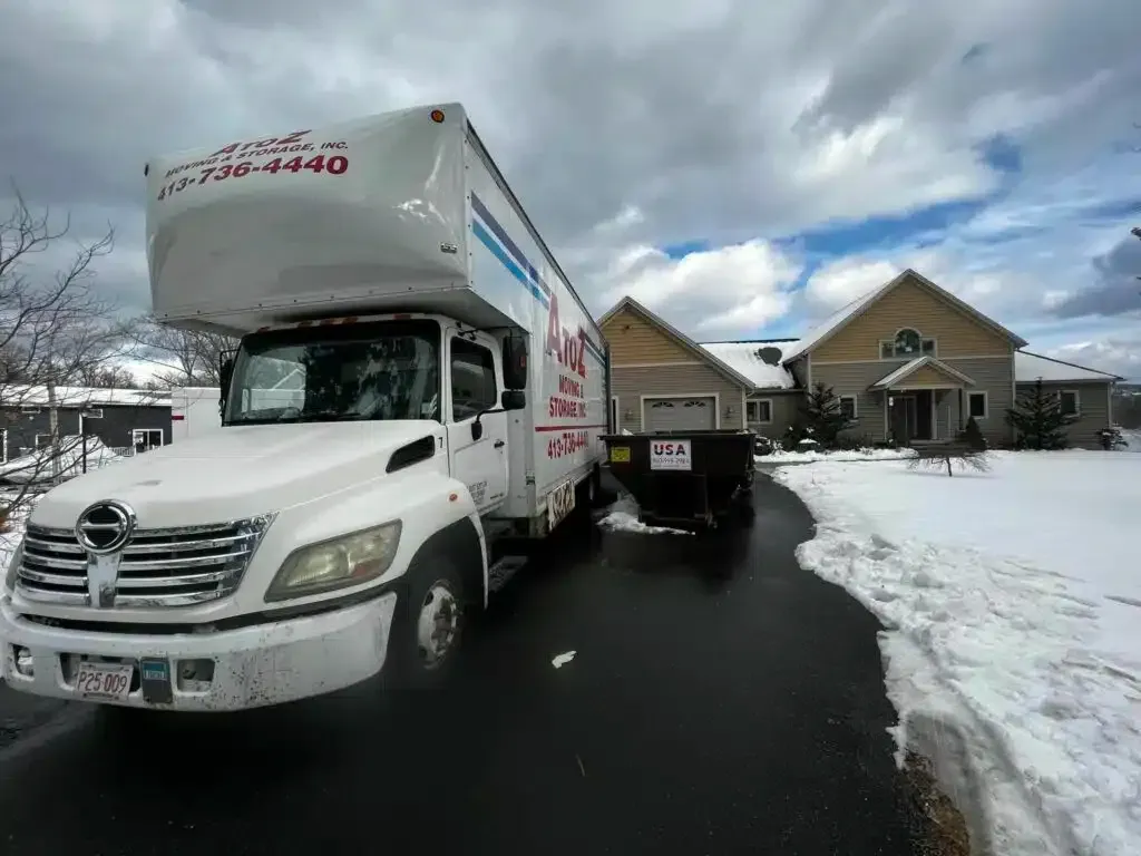 White moving truck and trailer on a snowy driveway in front of a house under cloudy skies
