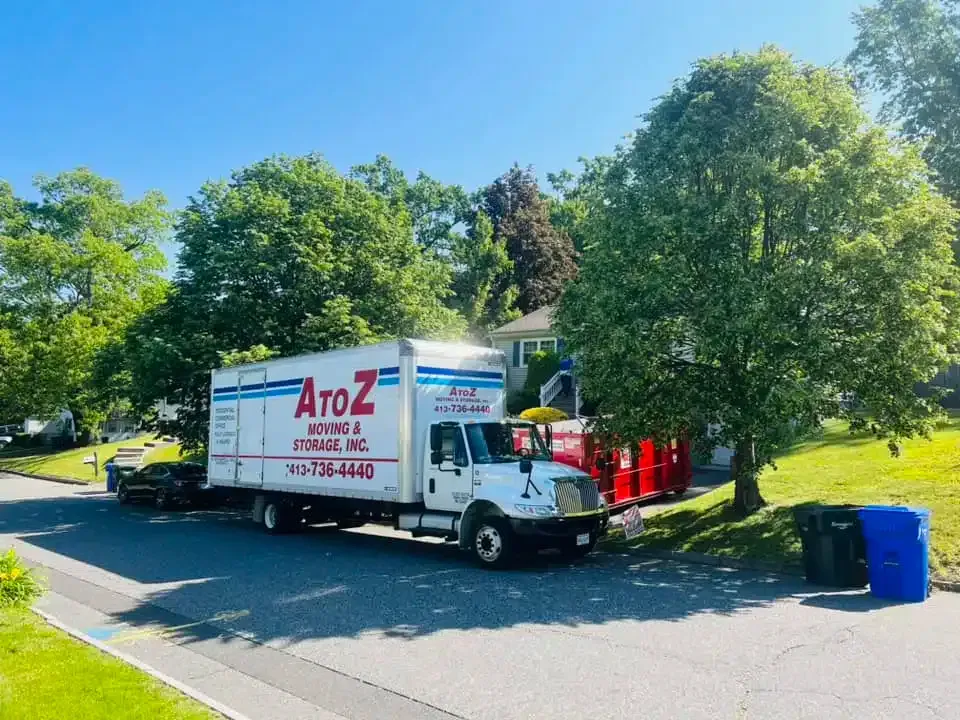 Moving truck parked on a tree-lined street beside blue recycling bins on a sunny day