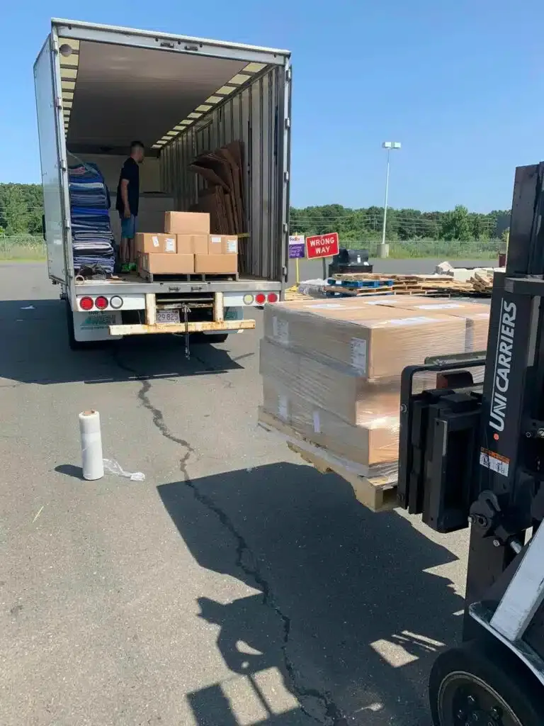 Truck being unloaded in a parking lot, with cardboard boxes on pallets and a forklift nearby.