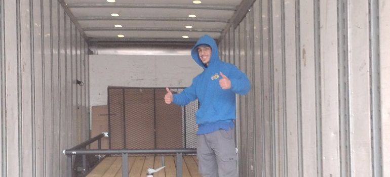 Person in a blue hoodie and gloves stands inside a shipping container near stacked cardboard boxes.