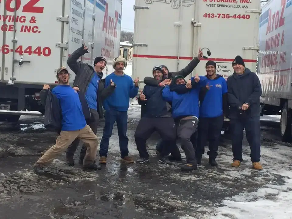 Six movers in blue shirts pose between two moving trucks on a snowy street.