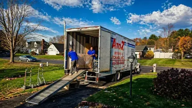 Workers loading furniture into a moving truck via a ramp outside a suburban house