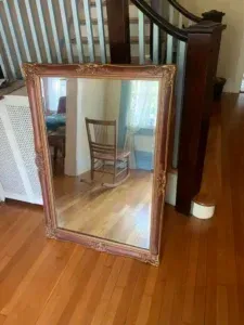 Framed mirror leaning on hardwood floor, reflecting a wooden chair and hallway stairs.