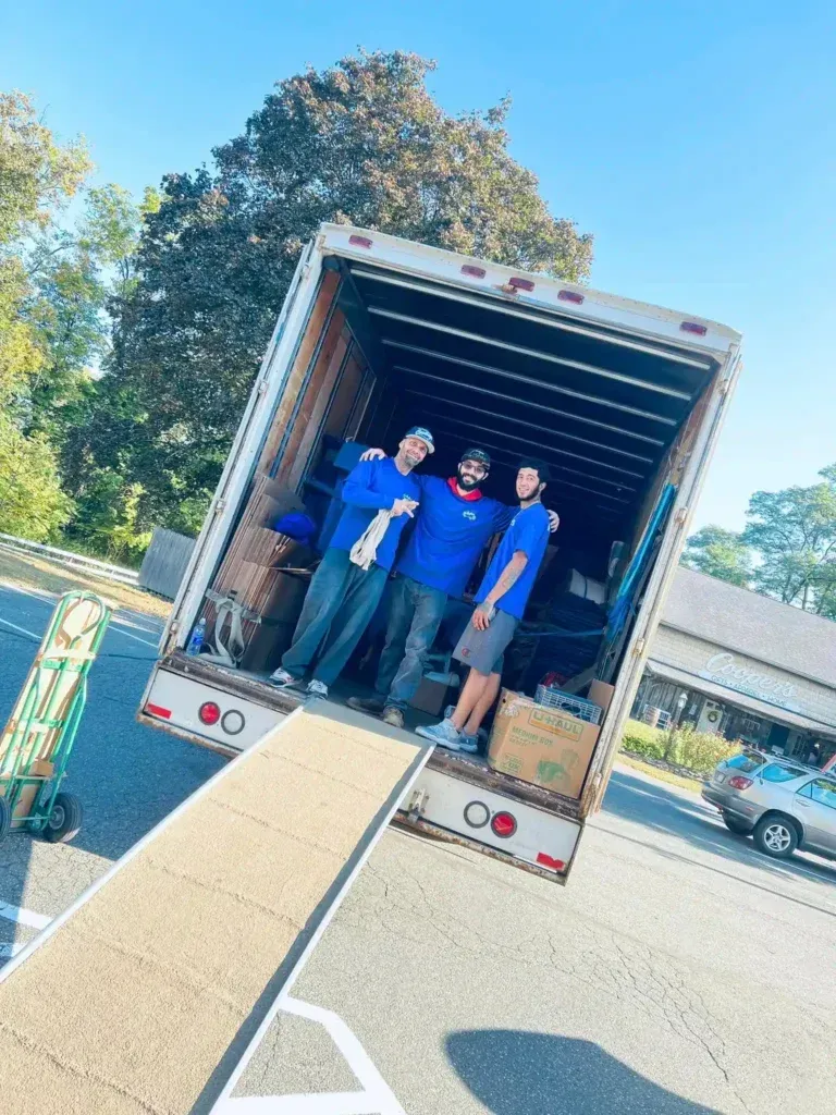 Three people in blue shirts stand in the back of a moving truck on a sunny street.