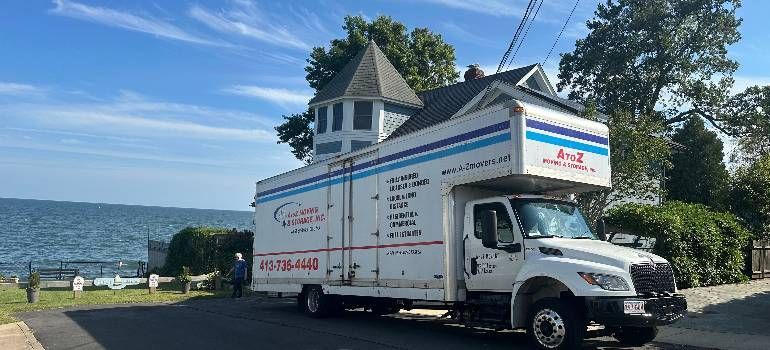 White moving truck parked beside a lakeside house with blue sky and water in the background