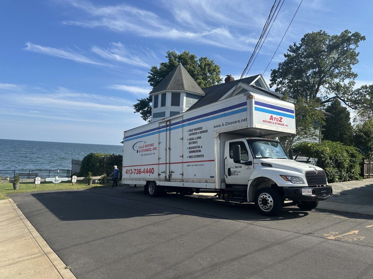 White moving truck parked on a sloped street near the waterfront, with houses and trees in the background