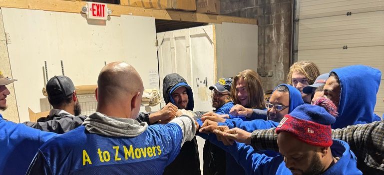 People in blue A to Z Movers shirts huddle together with hands stacked indoors.