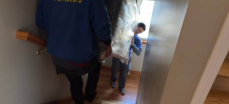 Two people walking down a narrow indoor staircase, one in a blue jacket and one in a light jacket.