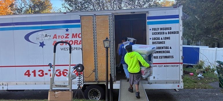 Two movers loading a wooden cabinet into a moving truck with open rear doors.