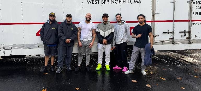 Six people posing outdoors in front of a truck in West Springfield, MA, one wearing neon green shoes