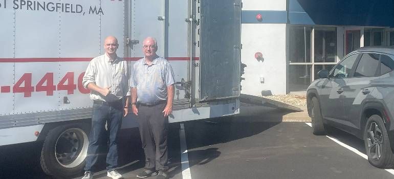 Two men standing beside a white moving truck on a sunny street outside a building