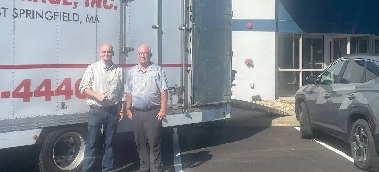 Two men stand beside a white box truck and a gray SUV outside a building on a sunny day.