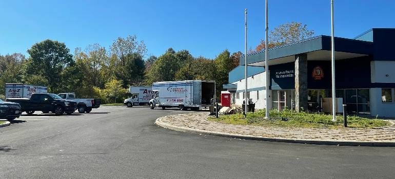 Parking lot outside a blue commercial building on a sunny day, with trucks and a white van near the entrance