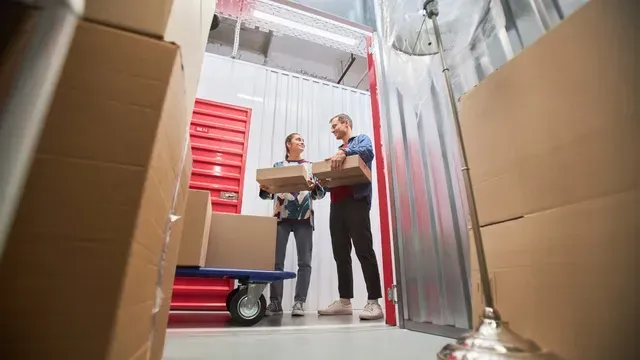 Two movers carrying boxes in a storage unit aisle beside a red dolly.
