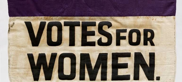 Vintage banner reading “VOTES FOR WOMEN” in black on cream fabric with a purple border