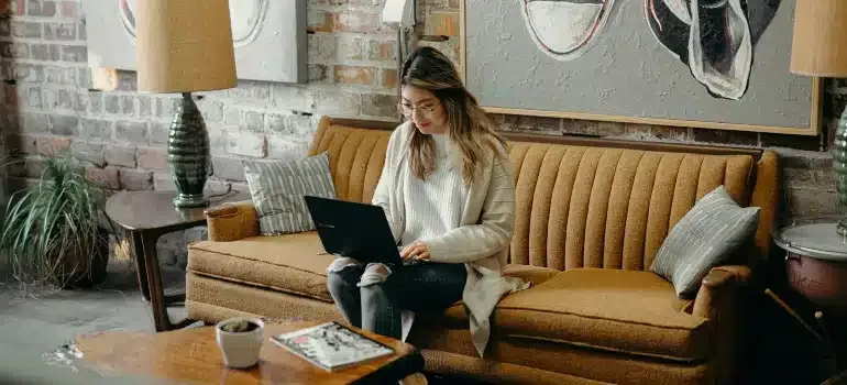 Woman using a laptop on a mustard sofa in a cozy living room with a lamp and brick wall.