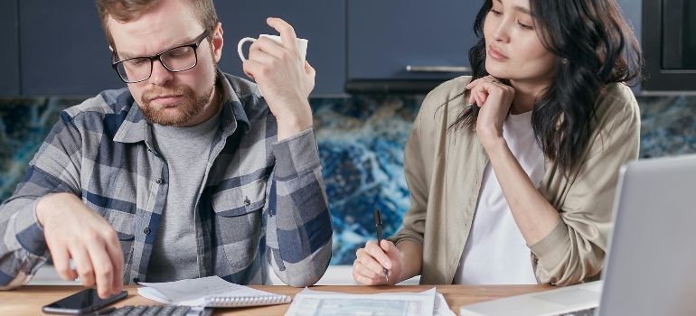 Two coworkers at a table, one frustrated with paperwork while the other looks on thoughtfully
