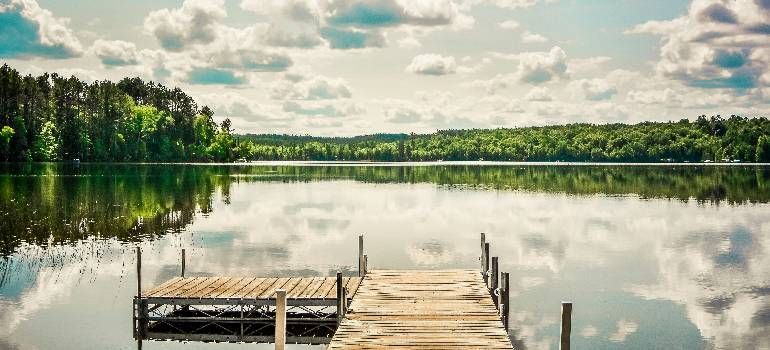 Wooden dock extending into a calm lake, reflecting cloudy sky and green forest shoreline.