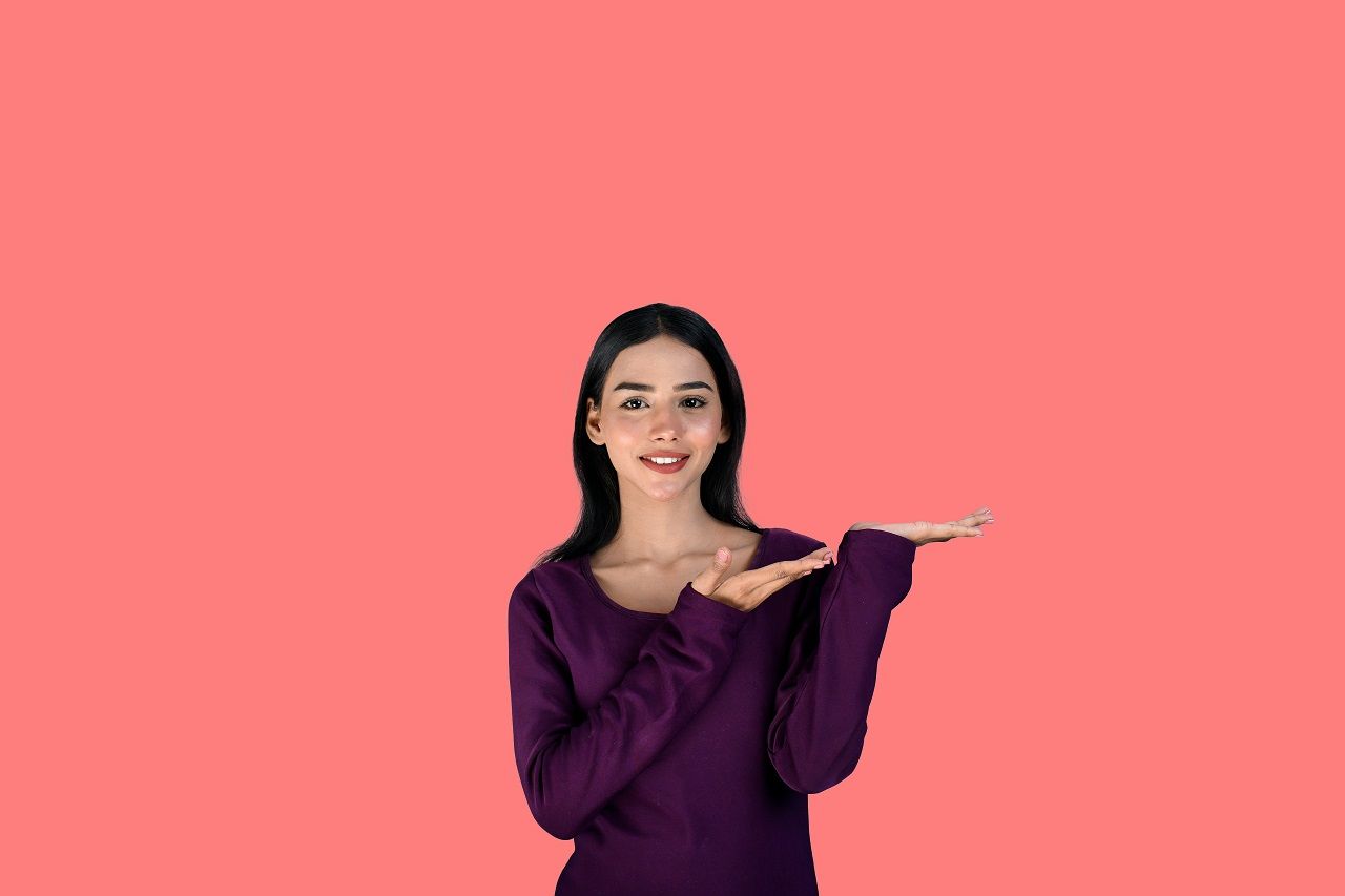Woman in a purple top gesturing with both hands against a pink background