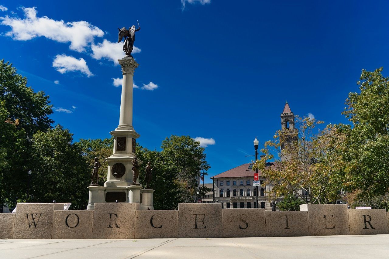 Stone monument in a sunny plaza with a tall column and eagle statue, framed by trees and buildings