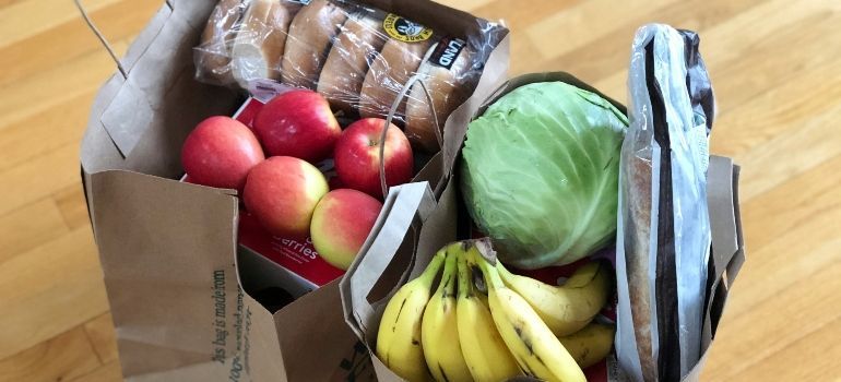 Open grocery bag with apples, bananas, cabbage, bread, and snack packs on a wooden floor