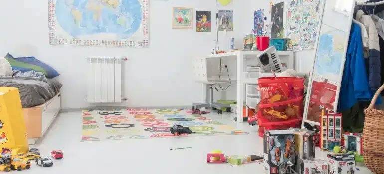 Child’s playroom with scattered toys, colorful rug, white shelves, maps on the wall, and a bed at left.