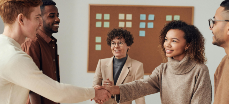 Four coworkers shaking hands in a warm office meeting room, smiling and facing each other.
