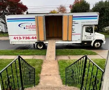 Moving truck with open side door and ramp extending to a sidewalk, flanked by black front steps.