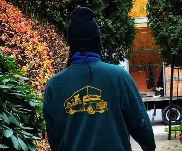 Person in a black beanie and teal jacket with yellow truck graphic, seen from behind in a garden setting