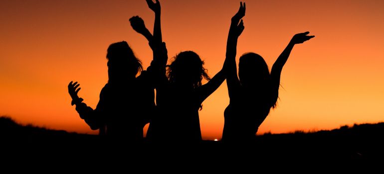 Silhouettes of three people dancing with raised arms against an orange sunset sky