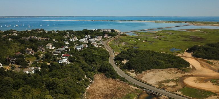 Coastal town with scattered houses, winding road, and green wetlands beside blue ocean under a clear sky