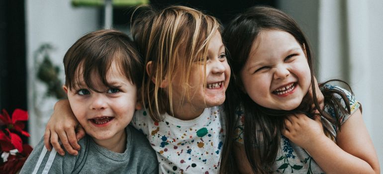 Three smiling children sitting close together indoors, with two looking at the camera and one looking aside