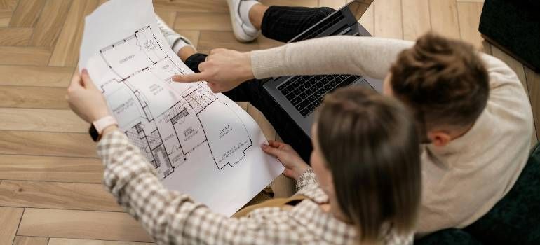 Two people reviewing printed floor plans together on a wooden table, pointing at the drawings.