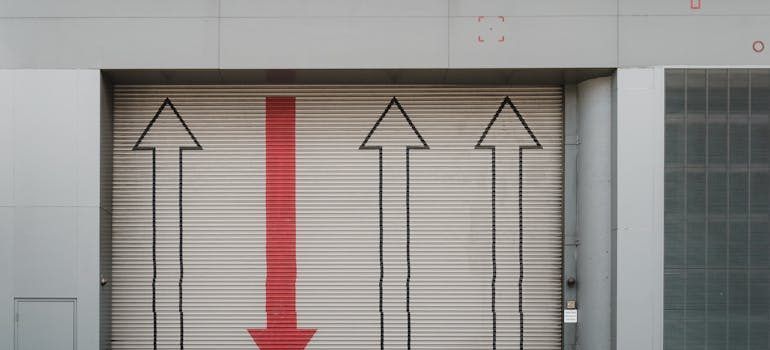 Closed metal door with black arrows and a red downward arrow in a tiled wall opening