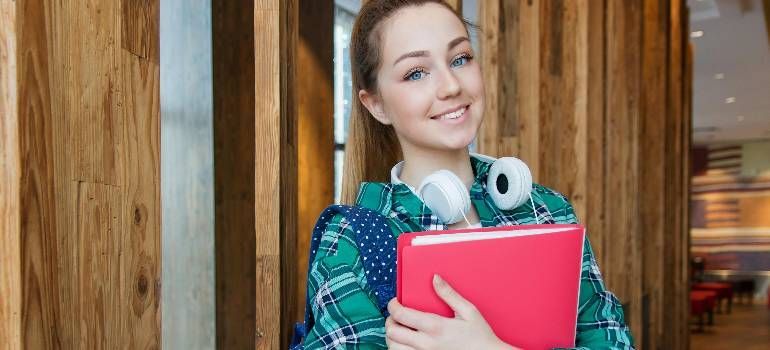Smiling student holding a pink notebook and headphones in a wood-paneled hallway